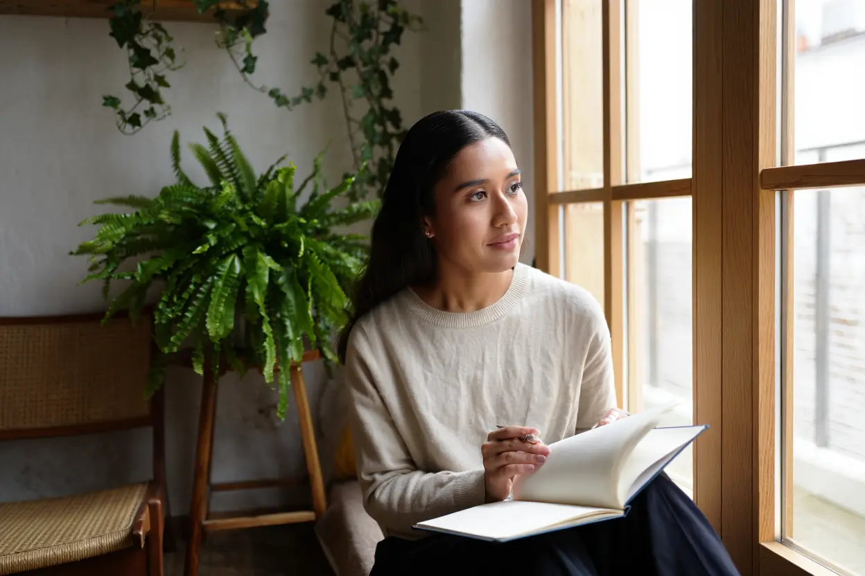 Femme assise près d’une fenêtre, écrivant dans un carnet, lumière naturelle et plantes vertes en arrière-plan. être soi-même citation naturelle dans le cadre.